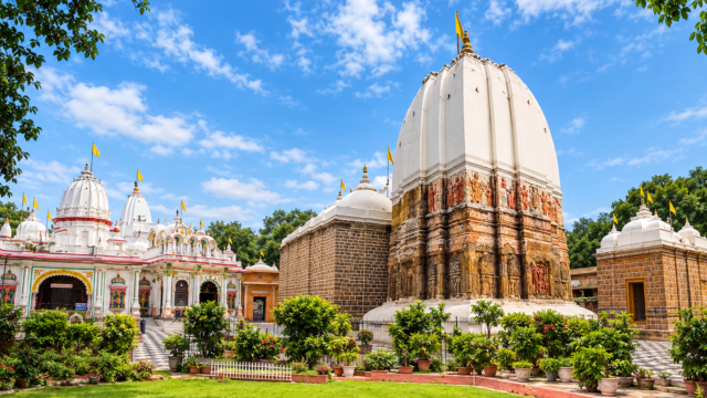 White Hindu temple with a tall dome, intricate stone carvings, colorful deity sculptures, and yellow flags against a bright blue sky.